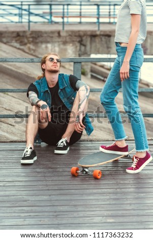 cropped image of boyfriend with tattoos sitting on bridge and girlfriend standing on skateboard