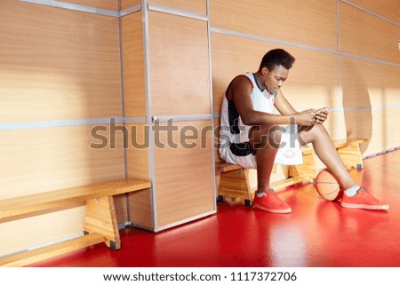 African-American man in basketball uniform sitting browsing smartphone and enjoying good music while sitting on bench in gym. 