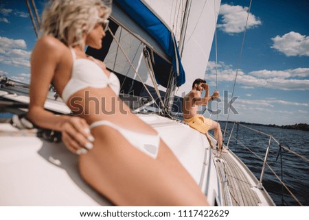 selective focus of woman in bikini having sunbath and her boyfriend sitting behind on yacht 