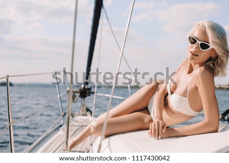 attractive young woman in sunglasses and bikini looking at camera on yacht 