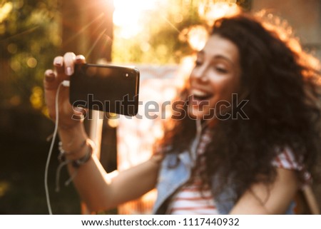 Image of cute young beautiful curly woman outdoors in park make selfie by mobile phone listening music with earphones.