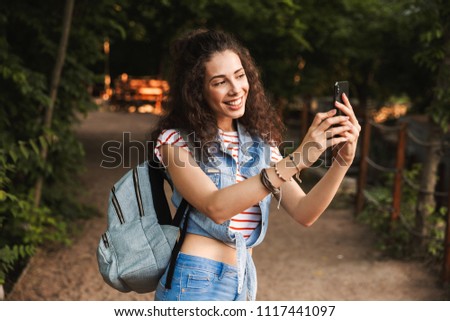 Photo of brunette pretty woman 18-20 with backpack smiling and photographing nature on smartphone while walking through green park