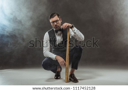 serious stylish man in eyeglasses crouching with baseball bat and looking at camera on black
