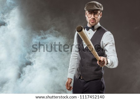 serious man in bow tie and cap holding baseball bat while standing in smoke 