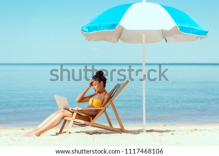 side view of smiling african american female freelancer sitting on deck chair and using laptop under beach umbrella in front of sea 