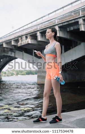 young sportswoman holding smartphone, earphones and sports bottle  