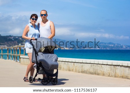 parenting and travel concept - happy young parents with baby stroller on the beach