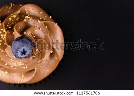Cupcake with whipped chocolate cream, decorated fresh blueberry, gold confectionery sprinkling on black background. Picture for a menu or a confectionery catalog. Top view. Close up.