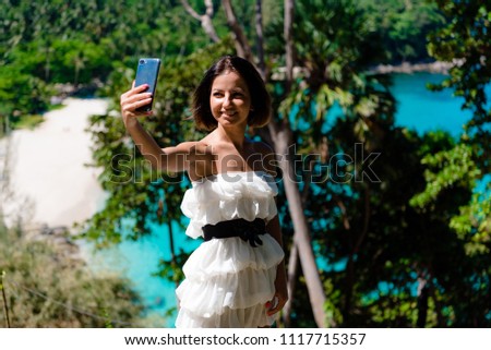 A young, attractive Girl takes Pictures of herself on a mobile phone camera, against the background of a tropical landscape.doing a selfie.