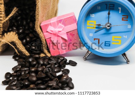 Coffee beans in a sack with blue alarm clock and pink gift box put together on a white surface.