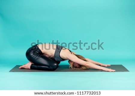 young and active blonde woman practicing yoga and meditating on a yoga mat in studio on blue background