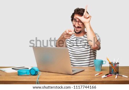 young bearded graphic designer with a laptop gesturing "loser" with fingers mockingly, with a happy, proud, and self-satisfied look.