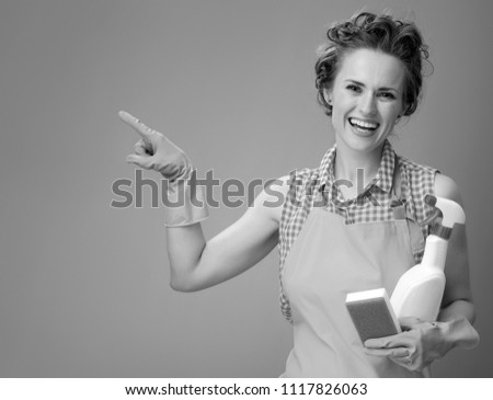 Big cleaning time. happy modern woman in a apron with kitchen sponge and a bottle of detergent pointing at something on background