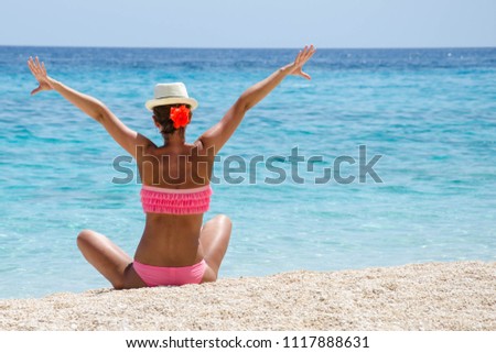 Joyful girl in pink swimwear sitting on a beach and watching turquose sea.