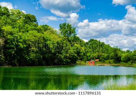 Rural swimming pond with change room, beach and dock along the far shore on a bright summer day with a cloudy blue sky.