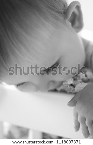 Black and white photograph of a Caucasian baby leaning on the edge of his bed with a sweet expression.
