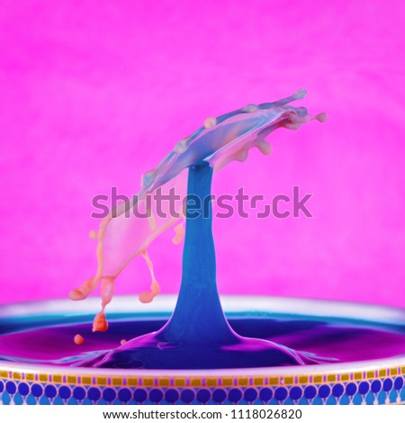 A colorful water drop with a blue spout and tumbling umbrella shape, on a hot pink background. Created with orange coloured milk dropped into blue liquid.