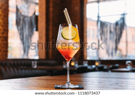 A colorful, cold drink. A white-orange drink in a glass with ice cubes, lime, silver straws and orange. The drink is on a napkin, a wooden table. In the background the interior of the bar.