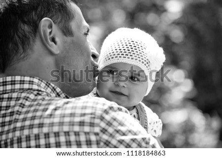 paternal care. Father kissing and holding small daughter baby girl cute hazel-eyed kid tiny little child wearing white flower beanie hat outdoor on sunny summer day on blurred green background