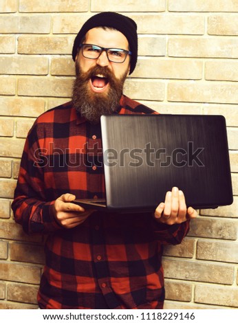 Bearded man, long beard. Brutal caucasian happy unshaven hipster holding laptop in red black checkered shirt with hat and glasses on beige brick wall studio background
