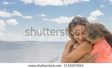 Little cute girl baby and young mother at beach. Mom sits on the shore, and her daughter is coming from behind. The kid hugs and kisses her mother. Mother and her child.
