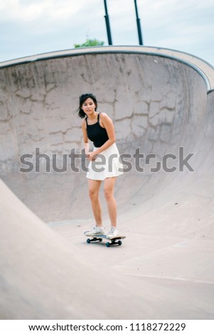 A young Chinese Asian teenage girl is skateboarding in a park during the day. She is wearing a skirt and black top and has her hair tied up. She is smiling as she skates.