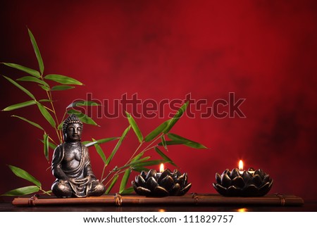 Buddha in meditation with burning candle and bamboo leaf on red background.