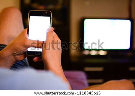 A man holds a smartphone in his hands using a DLNA in the living room in front of the TV, enjoying numerous content over the Internet. Shallow depth of field. Background. Natural light room. Royalty-Free Stock Photo #1118345615