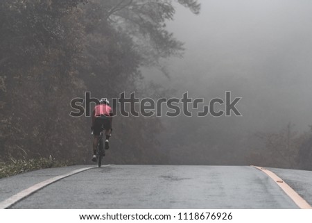Rear side of Asian cyclist in pink jersey ride his bicycle up high on hill in bad weather day. In cold, mist, raining and windy day he cycling to complete his mission. Royalty-Free Stock Photo #1118676926