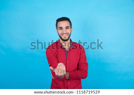 Handsome young man wearing a casual outfit, and he is calling someone to come, on his face big smile, standing on a blue background.