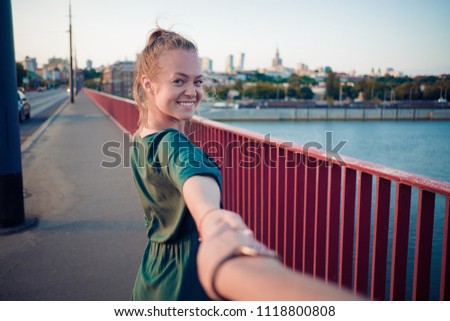 Follow me. Cheerful young woman holding hand and walking with her friend on the bridge