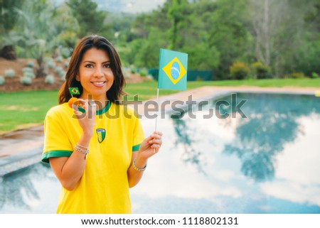 Smiling young woman (25-30) wearing a Brazilian national team jersey and eating a Brazilian "coxinha" chicken appetizer while waving a Brazilian flag with a pool reflection - soccer/football fan Royalty-Free Stock Photo #1118802131