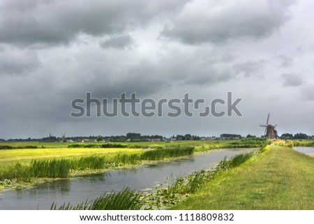 Typical Dutch landscape in summer with water and windmill under threatening weather conditions with heavy clouds in the sky, just before rainfall Royalty-Free Stock Photo #1118809832