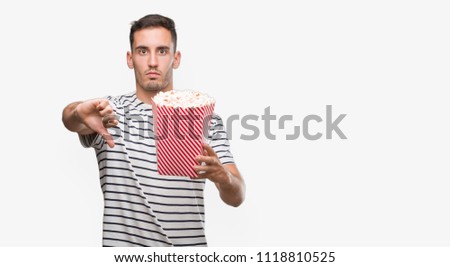 Handsome young man eating popcorn with angry face, negative sign showing dislike with thumbs down, rejection concept