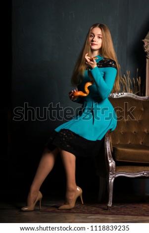 Beautiful young woman in deep sky blue dress sits on a chair with orange corn snake and looks at camera