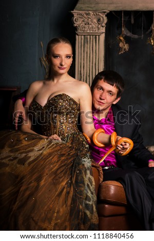 Beautiful couple in evening dress and dress suit sits with orange corn snakes, smiles and looks at camera