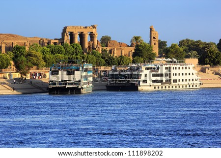 Egypt. Cruise ships docked at Kom Ombo on the Nile. The Temple of Sobek and Haroeris in background - seen colonnade of the Hypostyle Hall Royalty-Free Stock Photo #111898202