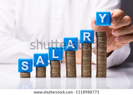 Person's Hand Making Salary Word By Placing Blue Cubic Blocks On Stacked Coins Over Desk Royalty-Free Stock Photo #1118988773