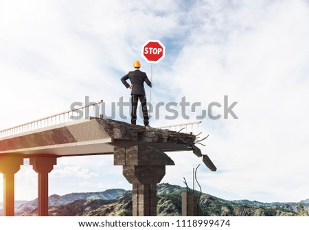 Rear view of engineer in helmet holding stop sign while standing on broken bridge with skyscape and sunlight on background. 3D rendering.