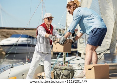 Senior yachtsman and his assistant loading boxes on boat before sailing trip on summer weekend Royalty-Free Stock Photo #1119201779