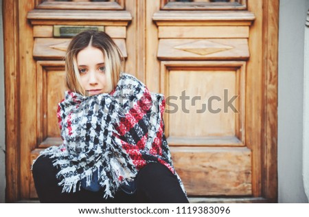 beautiful young blond woman sitting on the threshold of the house wrapped in a warm scarf, autumn