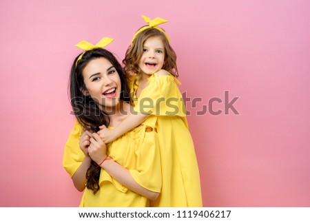 Crop view of positive brunette mother holding smilling daughter in same yellow dress looking at camera in studio on pink background