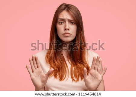 Displeased female model with freckled skin, raises eyebrows and frowns face, makes refusal gesture, says: No, I don`t want to to do it, leave me in pease, please! Discontent European woman indoor