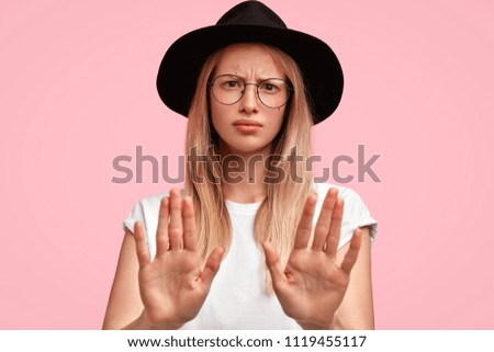 Photo of displeased blonde young female with discontent expression, keeps palms in front of camera, rejects something, doesn`t want to participate in fashion show, isolated over pink background