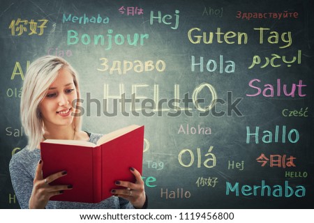Pretty girl holding a red book in front of a huge blackboard written with the word hello in different languages and colors. Opportunity for learning many languages for students.
