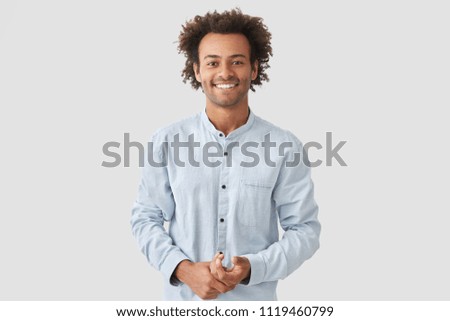 Horizontal shot of happy mixed race male with shining smile, feels glad as recieves bonus and praise for good work, has specific appearance, Afro hairstyle, dressed in fashionable white shirt Royalty-Free Stock Photo #1119460799
