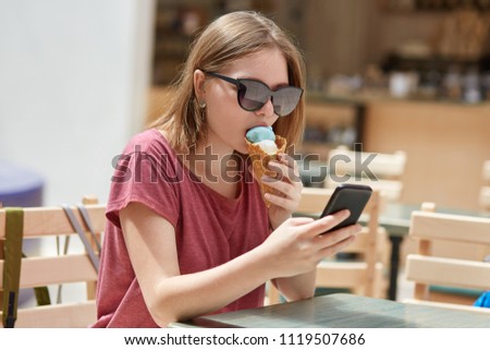 Shot of pleasant looking stylish young female hipster reads information on cell phone, surfes social networks, dressed in casual t shirt, eats delicious ice cream, enjoys spare time in summer