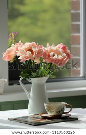 Gorgeous bouquet of peonies on enamel pitcher, ceramic tea cup on tablecloth on window background, warm summer day in garden, vintage style, vertical photo, daylight