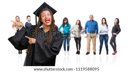 Young graduated black woman wearing braids pointing to the side, smiling surprised presenting something, natural and casual