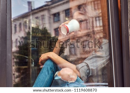 Young redhead curly girl wearing casual clothes and glasses sitting on a window sill with a takeaway coffee.